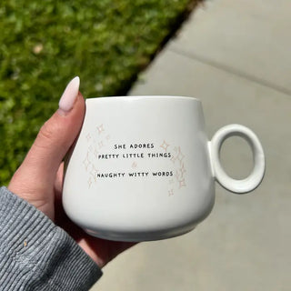 A hand with long nude nails holds a white ceramic mug with a loop handle outdoors. The mug has black text reading 'SHE ADORES PRETTY LITTLE THINGS NAUGHTY WITTY WORDS' with small gold starburst accents.