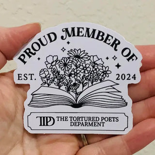 A hand holding a die-cut sticker that reads 'PROUD MEMBER OF EST. 2024' with an illustration of an open book and flowers.