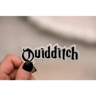 A hand with black nail polish holds a die-cut sticker featuring the word 'Quidditch' in a black gothic font with a white outline, set against a blurred beige background.