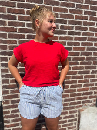 Person wearing a red t-shirt and blue striped shorts standing against a brick wall.