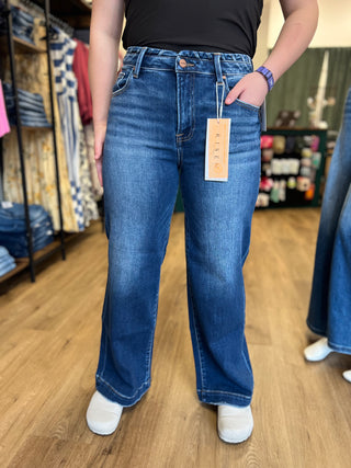 A person wearing medium-wash blue wide-leg jeans with a visible hangtag, standing in a clothing store on a wood floor.