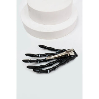 A black skeletal hand hair claw clip with a silver metal spring, displayed on a light gray surface with a white cylindrical prop in the background.