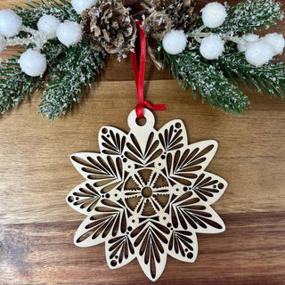 A laser-cut wooden snowflake ornament with a red hanging ribbon, displayed on a wooden surface with festive greenery and white pom-poms above it.