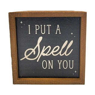 A square wooden sign with a dark charcoal face and natural brown wood frame. The sign features the phrase 'I PUT A Spell ON YOU' in cream-colored lettering, with 'Spell' in a cursive script and 'I PUT A' and 'ON YOU' in a serif typeface, accented with small cream stars.