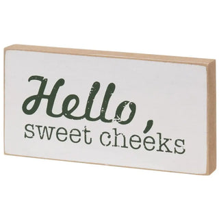 A rectangular wooden sign with a white face and natural wood edges, featuring the phrase 'Hello, sweet cheeks' in dark green lettering, shown on a plain white background.