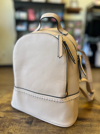 A light beige leather backpack with black trim, top handle, and zipper pulls, resting on a wooden table in a blurred indoor setting.
