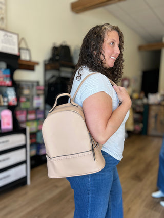 A woman with curly brown hair wearing a light blue striped t-shirt and blue jeans carries a beige faux-leather backpack with black trim in a retail store setting.