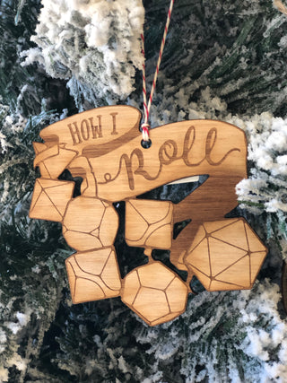 A wooden Christmas ornament shaped like a d20 die and other polyhedral game pieces, with the words 'How I feel' engraved, hanging from a string against a snowy tree branch background.