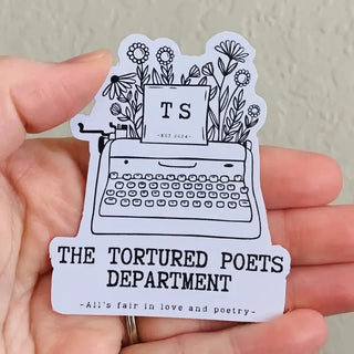 A hand holding a die-cut sticker featuring a black line art illustration of a typewriter with flowers and a paper insert reading 'TS -EST 2024'. Below the illustration, the text reads 'THE TORTURED POETS DEPARTMENT' and '-All's fair in love and poetry-'.
