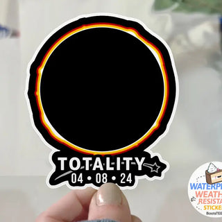 Sticker with 'TOTALITY' and date held by a hand on a blurred background