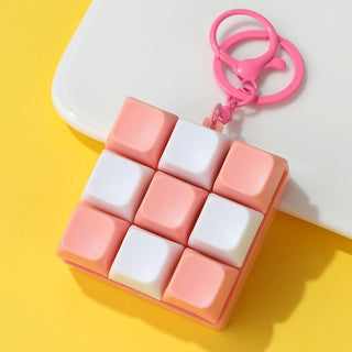 A square keychain with a 3x3 cube puzzle design in pastel pink and white, attached to a bright pink split ring and clasp, photographed on a yellow and white background.