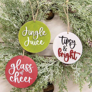Three round wooden hanging ornaments with holiday text, displayed on a frosted green pine branch. One ornament is light green and reads 'Jingle Juice' in white script.